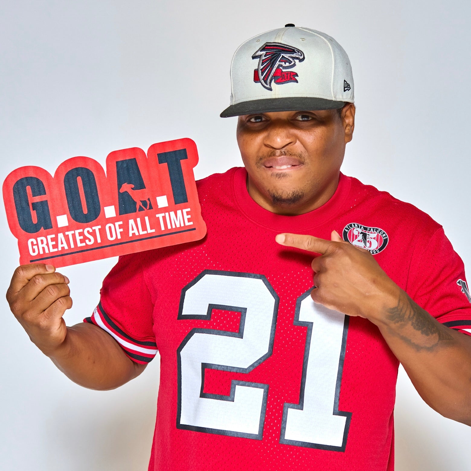Man in red Falcons jersey holding and pointing at a red “G.O.A.T Greatest of All Time” photo booth sign.